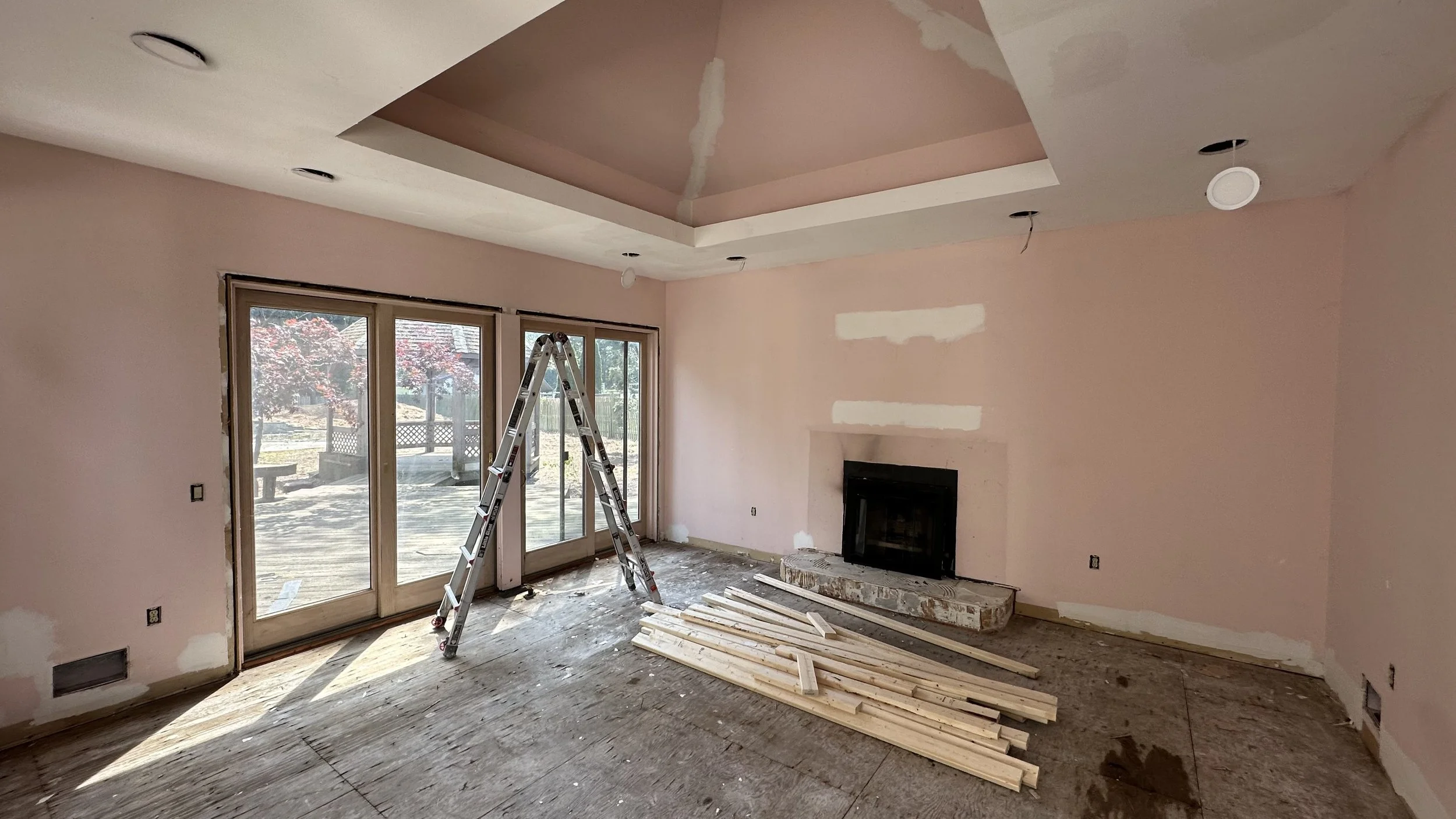 Living room under renovation with pink drywall, a ladder, wooden planks, and a fireplace, with large sliding glass doors leading outside.