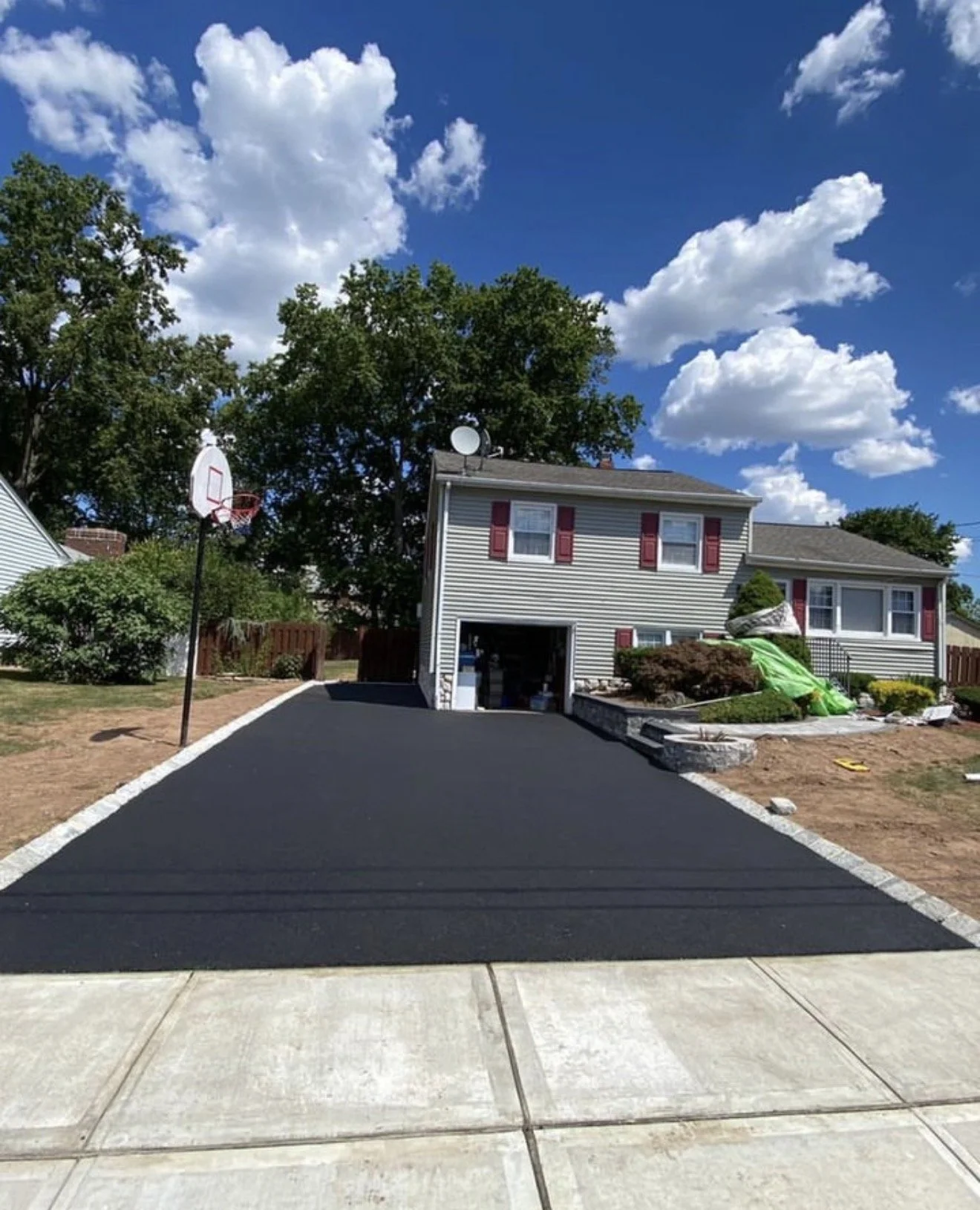 A newly paved black asphalt driveway leading to a two-story house with gray siding and red shutters, under a blue sky with white clouds.