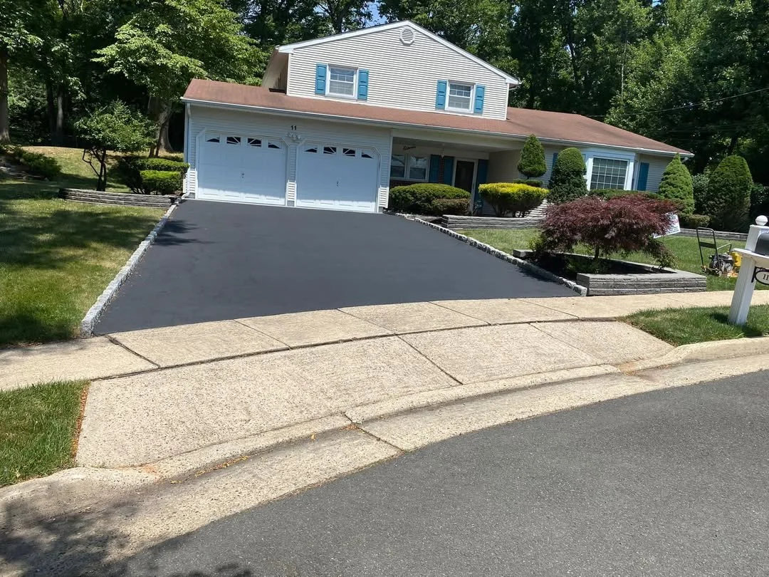 A house with a brown roof, white siding, and blue shutters, with a freshly paved black asphalt driveway leading to a two-car garage. The front yard has green grass, neatly trimmed bushes, and a small tree with purple leaves.