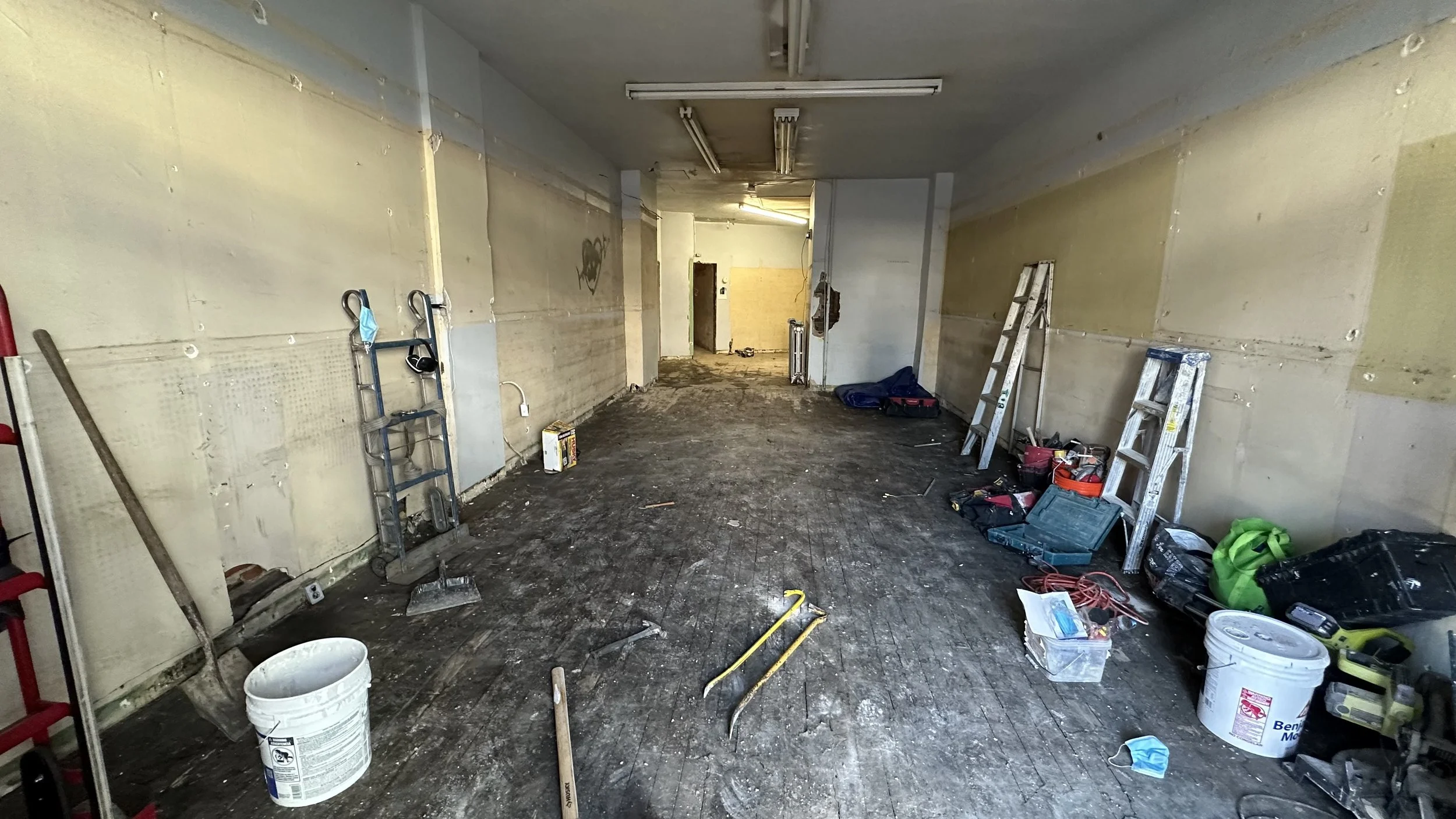 Empty room undergoing renovation with tools and materials scattered, ladders, a bucket, mask, and construction equipment.