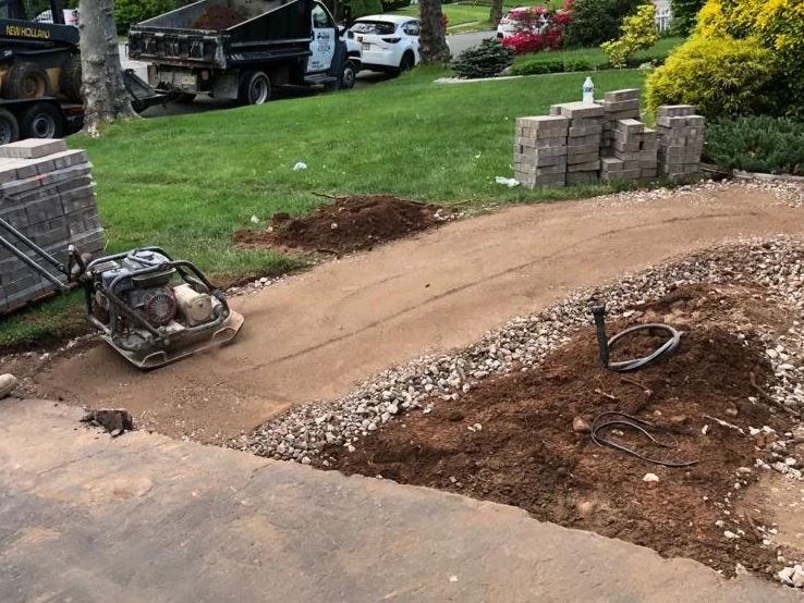 A pathway under construction with a dirt and gravel surface, a compactor machine on the pathway, and bricks stacked nearby. In the background, there are parked cars, trees, bushes, and a water bottle on a post.