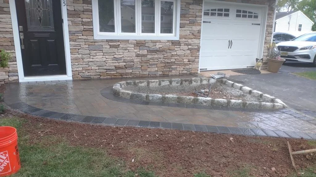 Front yard patio with newly laid concrete and stone border, adjacent to a house with stone and siding exterior, and a garage with a closed door.