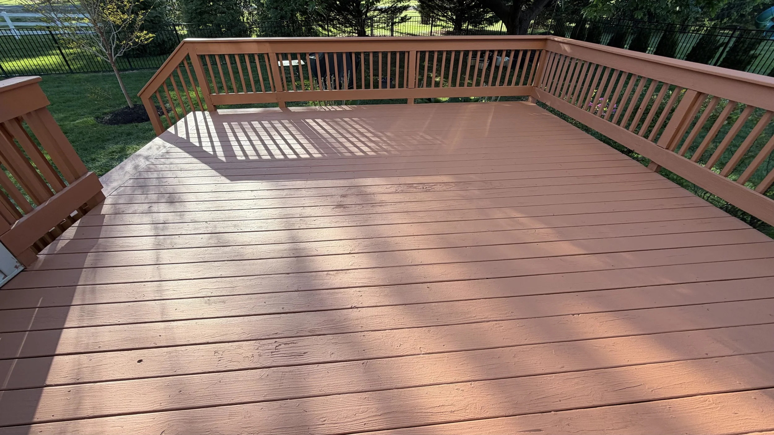 Photo of a wooden deck with a railing, shadows cast by the sun, and a backyard with grass, trees, and a black fence.