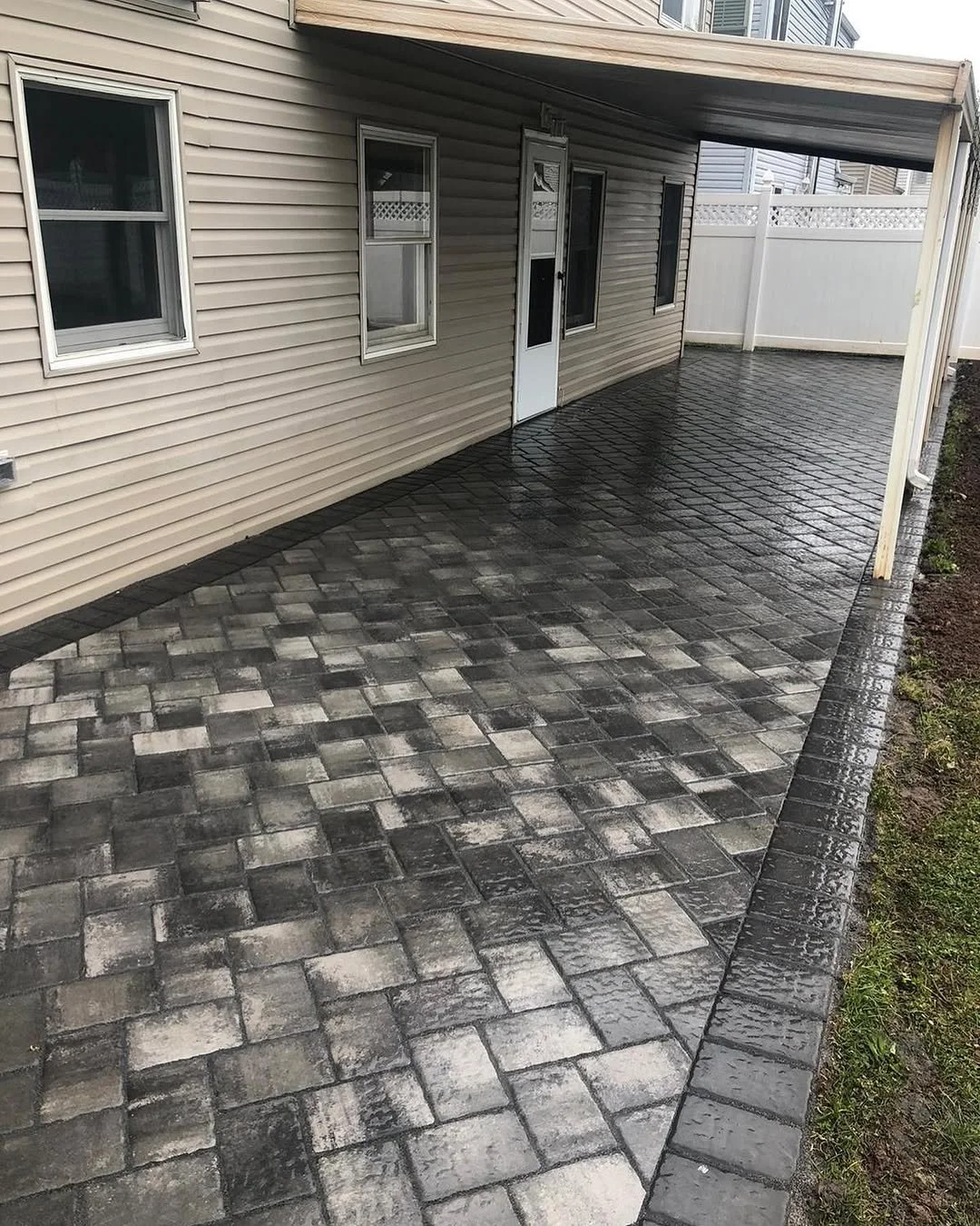 A newly paved outdoor patio with stone tiles in front of a house with beige siding, three windows, and a door. There is a covered section supported by wooden beams and a white fence at the back.