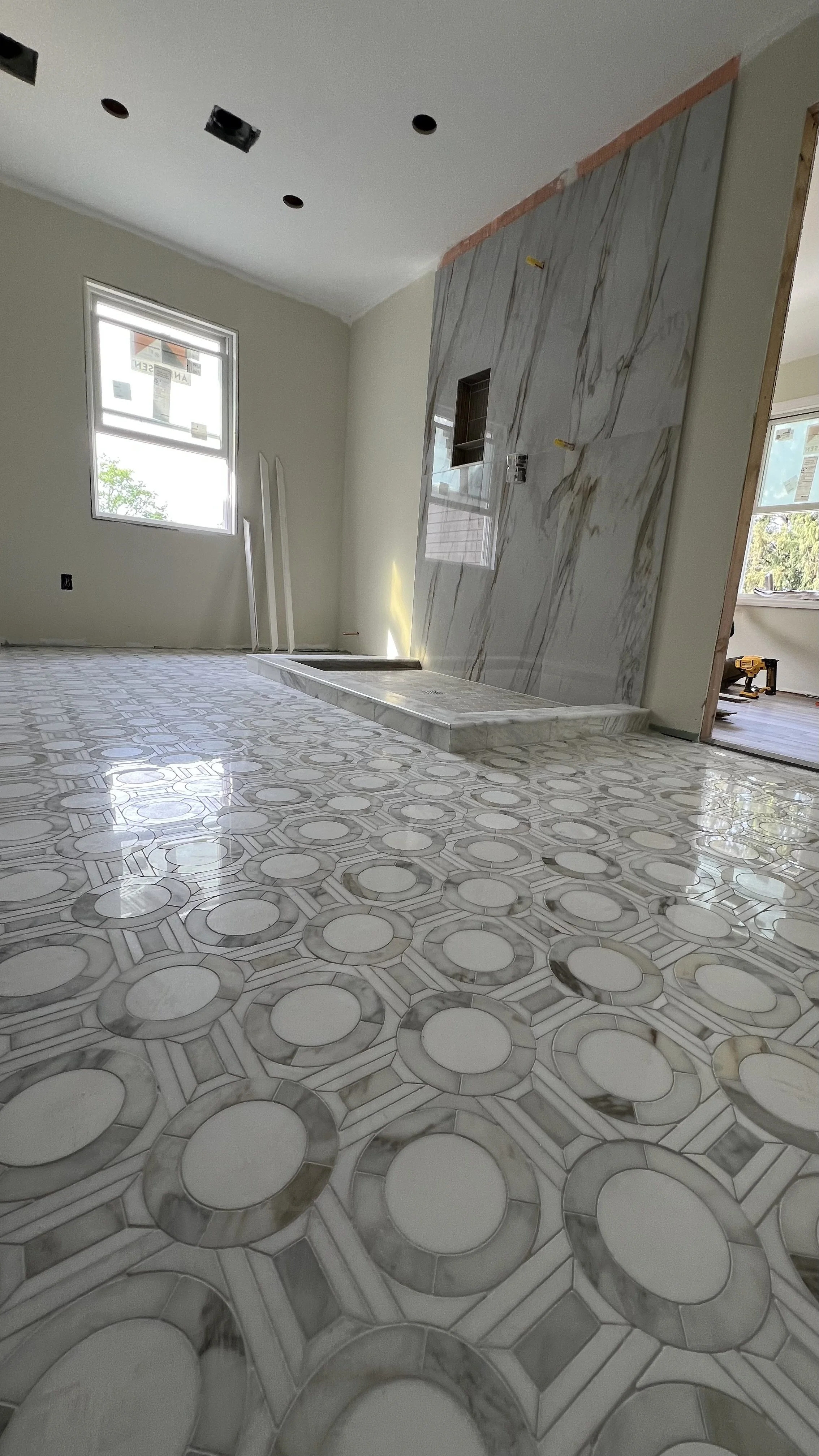 Room under construction with marble flooring, a wall with marble tiles, and a small platform or fireplace. There are windows and construction tools and materials visible.