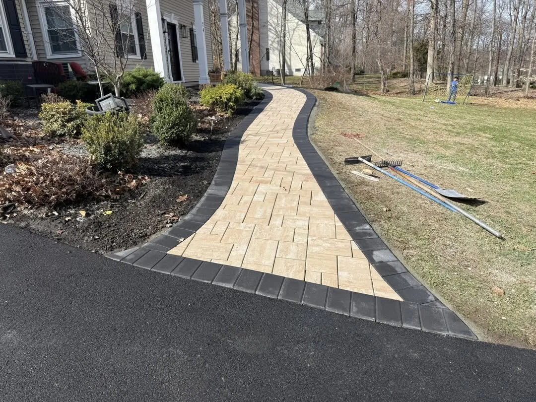 Newly paved winding walkway with beige pavers, black border stones, and edging bricks, leading away from a house with landscaping and garden tools on the grass.