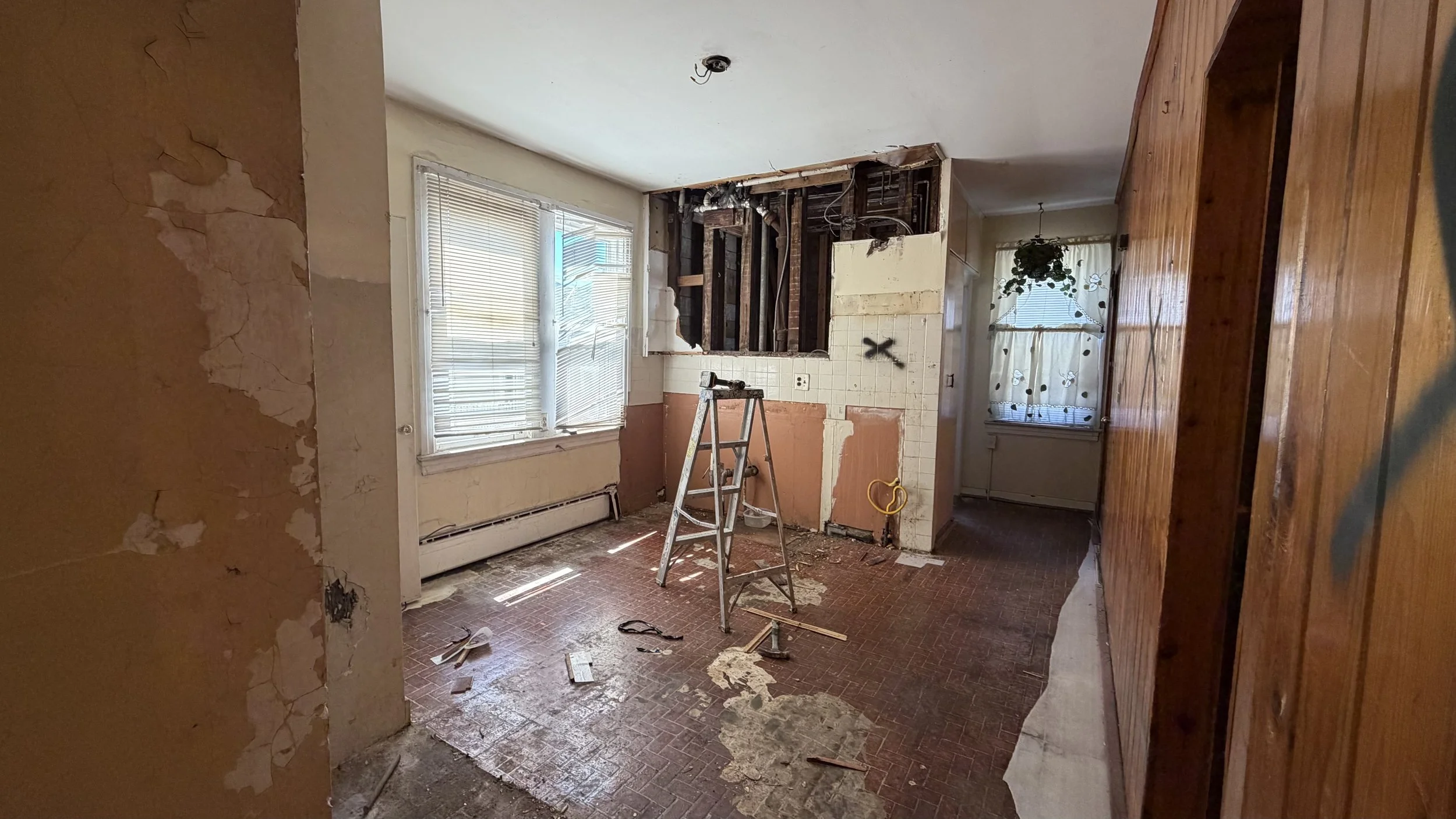 Room under renovation with exposed wall and ceiling, a ladder, and construction debris on the floor, with two windows, one covered with blinds and the other with curtains, and a partial view of a wooden wall.