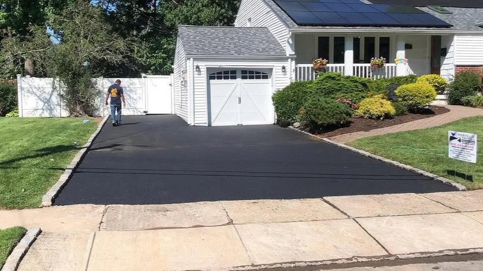 A man walking on a freshly paved black asphalt driveway leading to a white house with a garage door, surrounded by green bushes and grass, with solar panels on the roof, and a sign on the lawn.