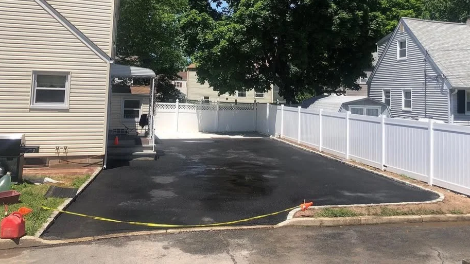 A freshly paved black asphalt driveway in front of a house, bordered by a white vinyl fence, with a tree providing shade in the background.