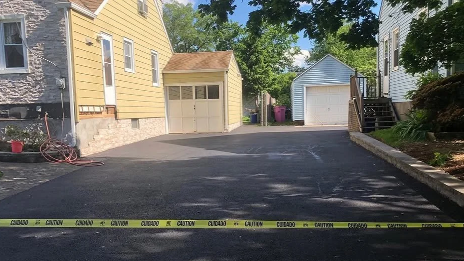 Freshly paved driveway with caution tape across it, yellow house and detached garages in the background, greenery and trees surrounding the area.