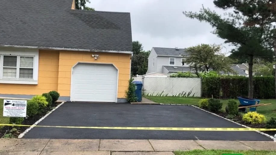Newly paved driveway with yellow caution tape in front of a yellow house with grey roof. The house has a single garage door, a window with white trim, and a blue recycling bin on the side. There is a garden with bushes, a wheelbarrow, and a white fen