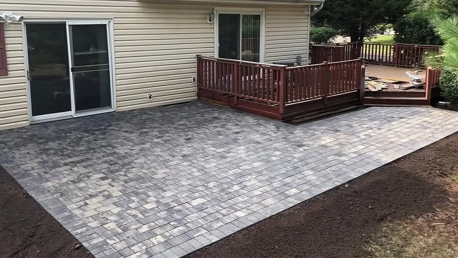 Backyard with a newly paved patio area, a wooden deck, a sliding glass door, and an outdoor space surrounded by trees and dirt.