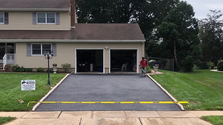 New asphalt driveway being installed in front of a suburban house with two garage doors, lawn, and trees. Caution tape is across the driveway and a worker is present.