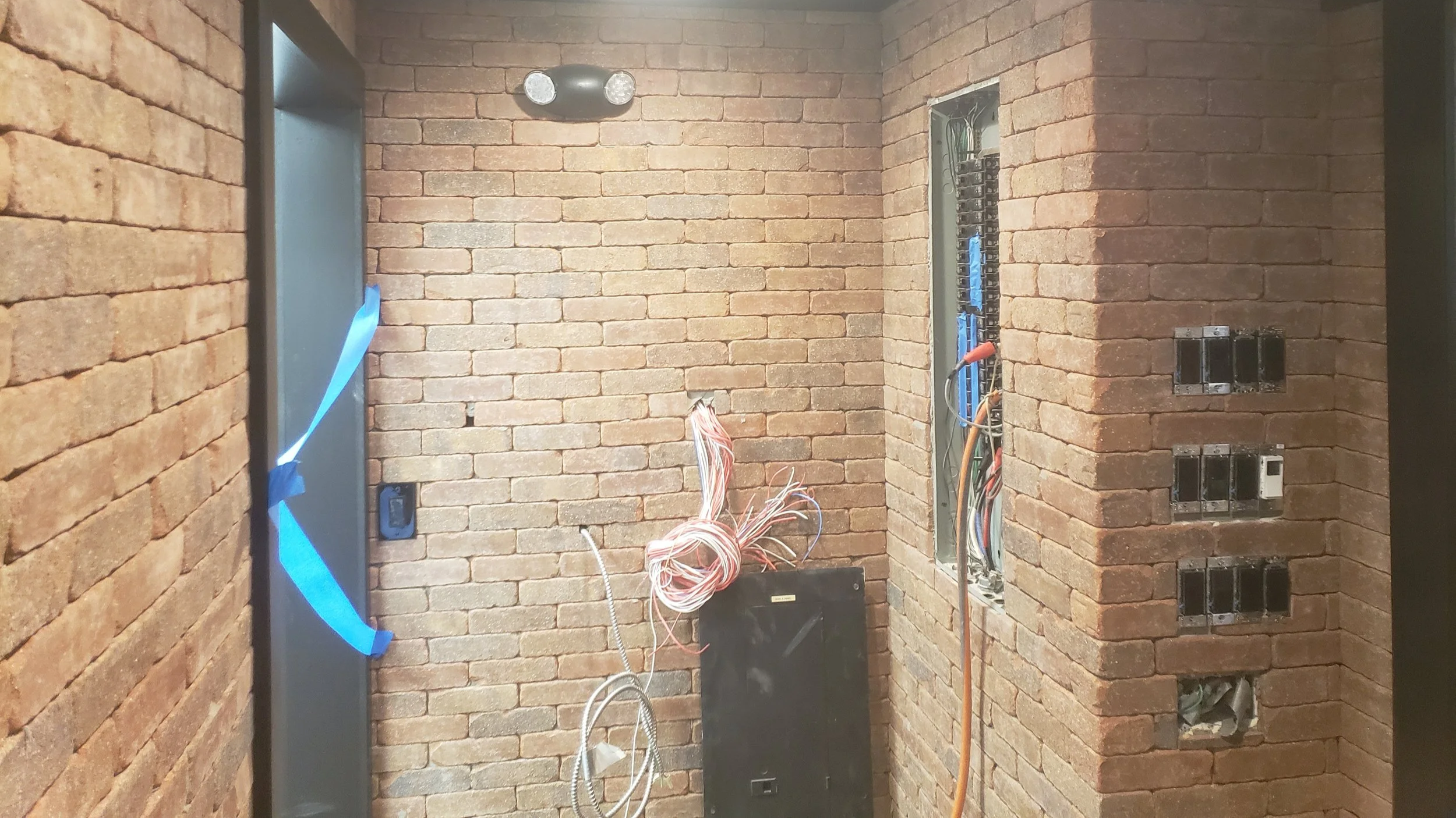Brick-walled room under construction or renovation with exposed wiring, electrical outlets, and switches, with a doorway covered with blue tape.