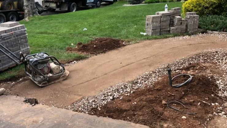 A construction site with a concrete roller, dirt, and gravel, with some stacked bricks and parked vehicles in the background.