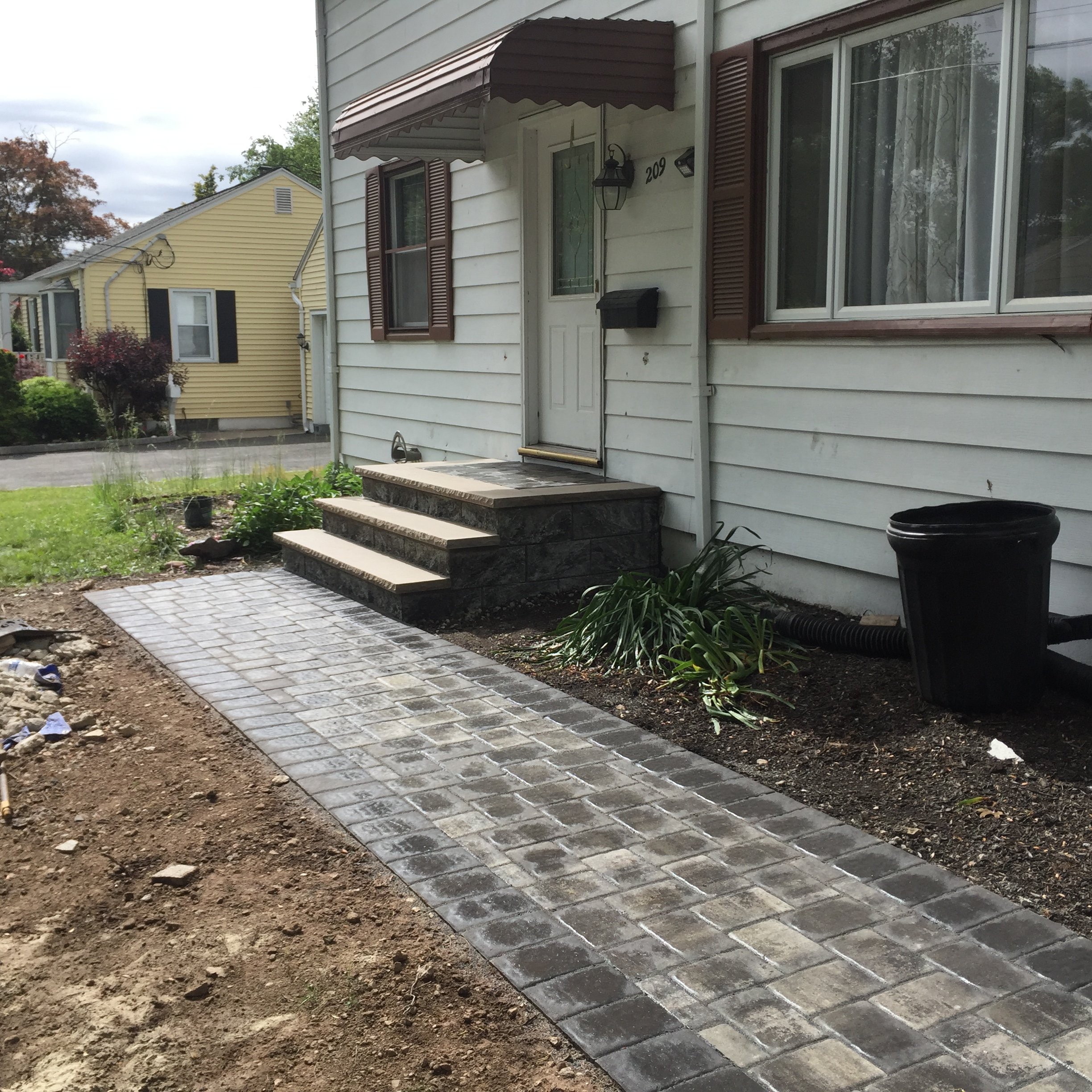 Front porch with steps and brick pathway leading to a white house with brown shutters.