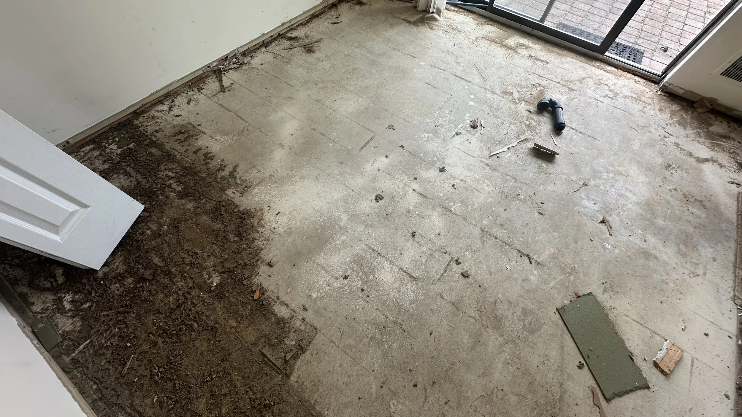 Room with partially removed flooring, exposing subfloor and construction debris, with door and sliding glass door leading outside.