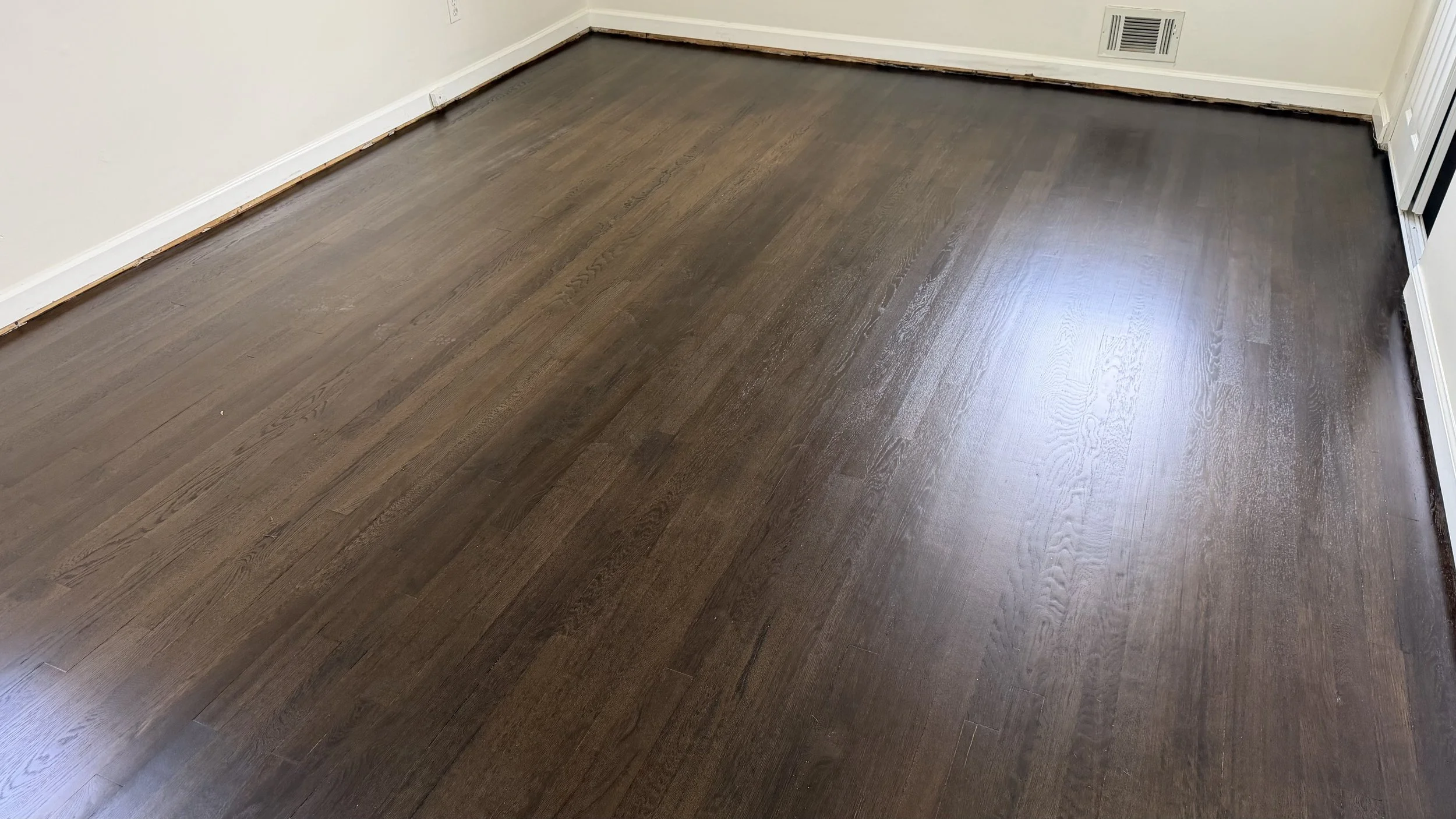 Finished dark hardwood flooring in a room with off-white walls, white baseboards, and a vent in the wall.