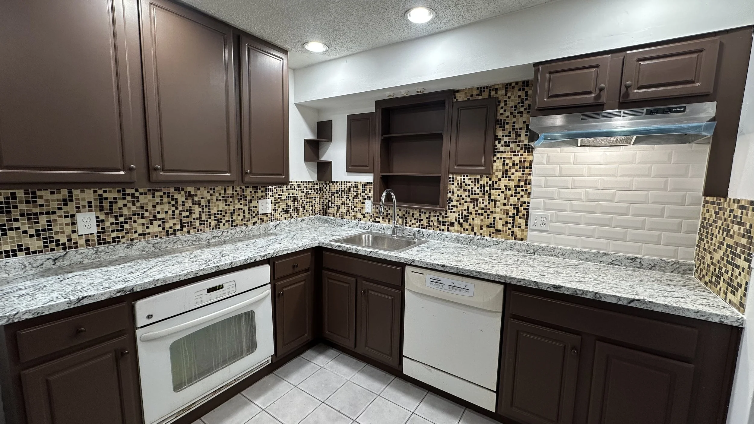 Kitchen with brown cabinets, granite countertops, mosaic tile backsplash, stainless steel sink, white appliances, and under-cabinet range hood.