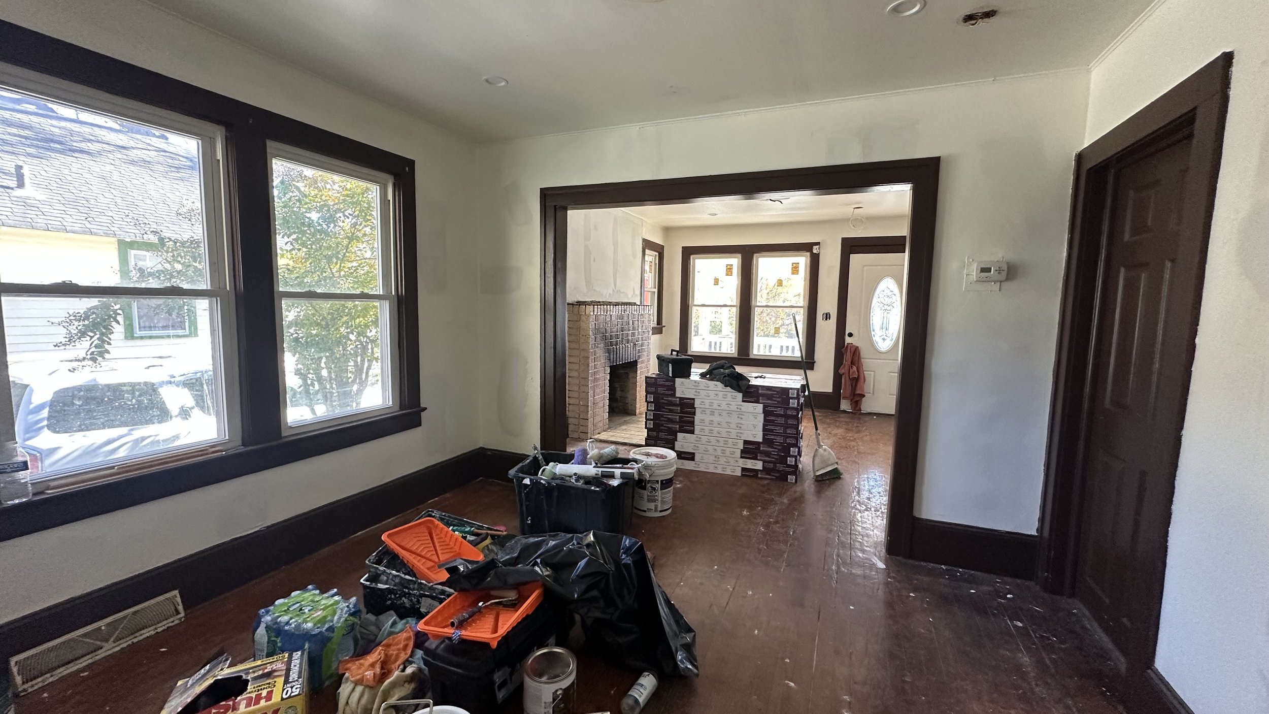 Interior of a house under renovation with tools, paint cans, and construction materials scattered on the wooden floor, white walls, and dark window and door frames.