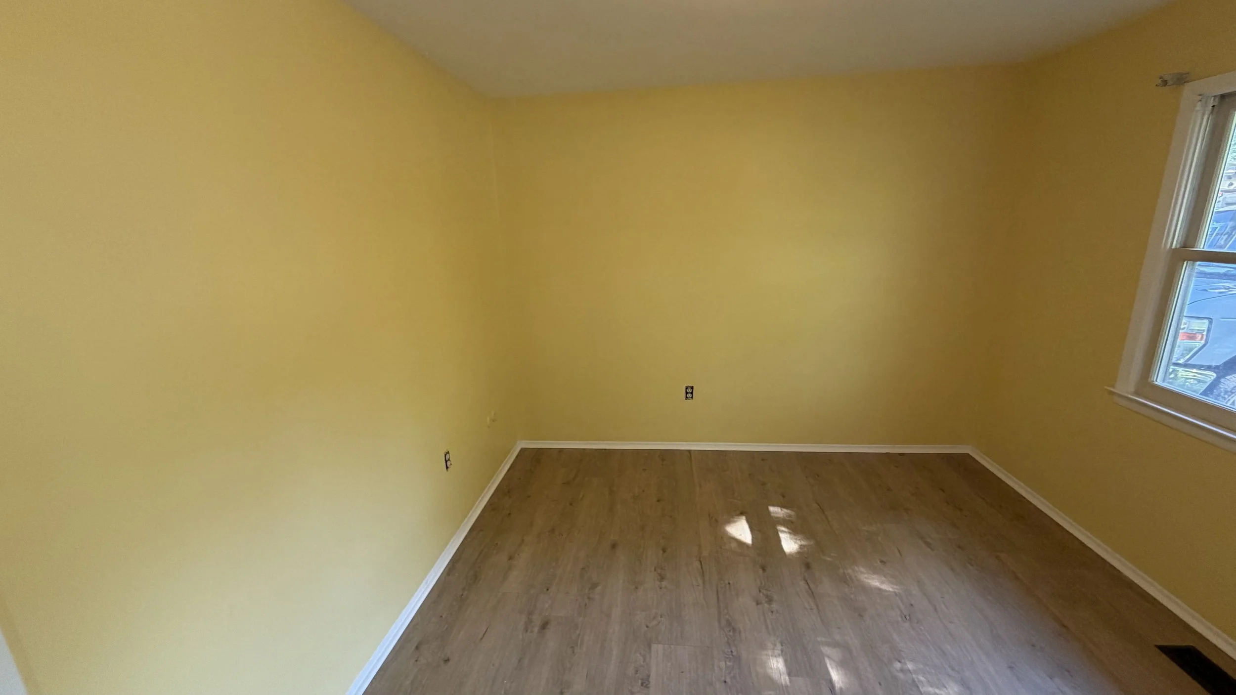 Empty room with yellow walls, hardwood floors, and a window on the right wall with sunlight streaming in.