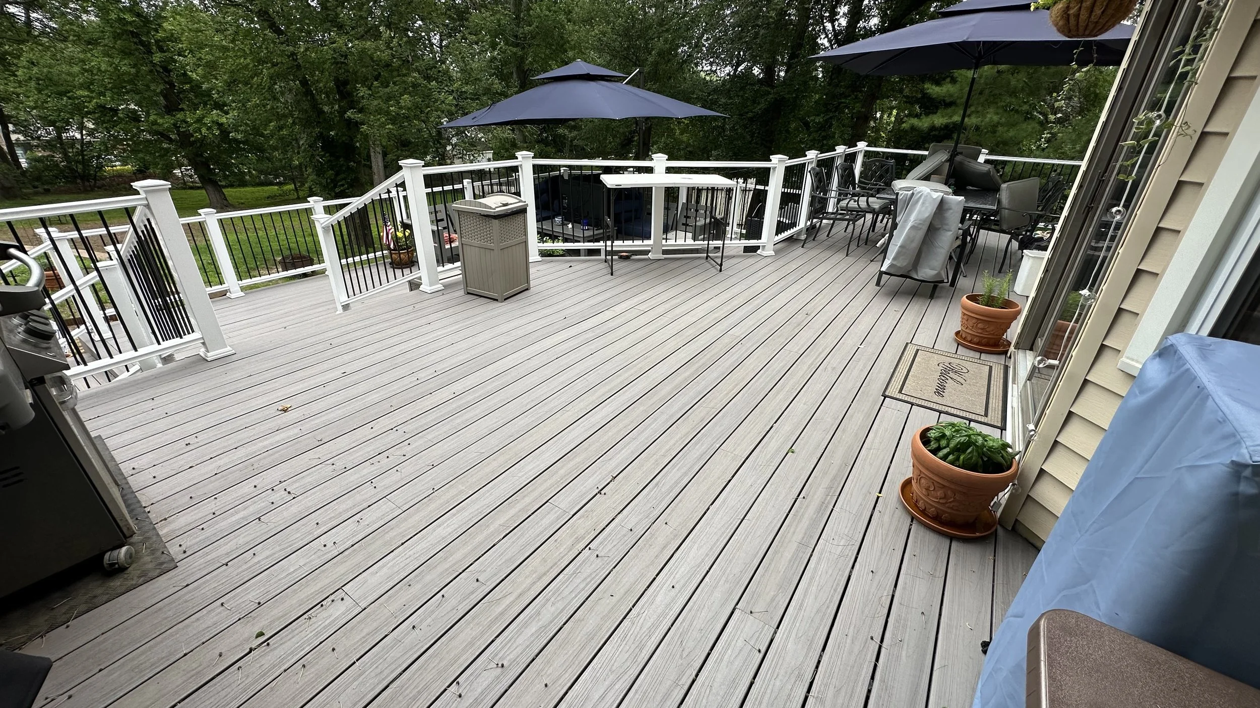 A spacious wooden deck with outdoor furniture, umbrellas, potted plants, and a view of green trees in the background.