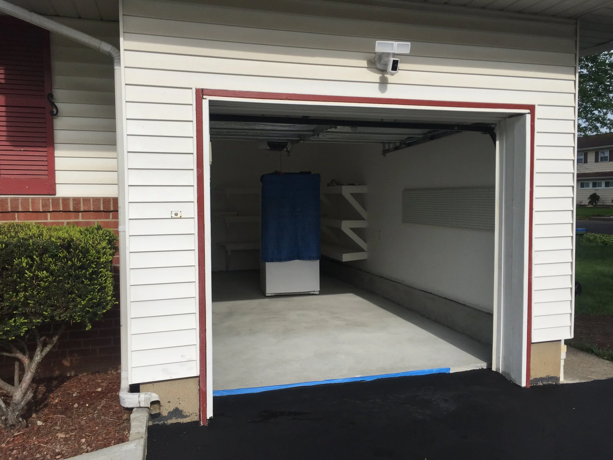 Empty garage with white walls, a blue-covered appliance, and white shelves inside, outside house with red brick and beige siding.