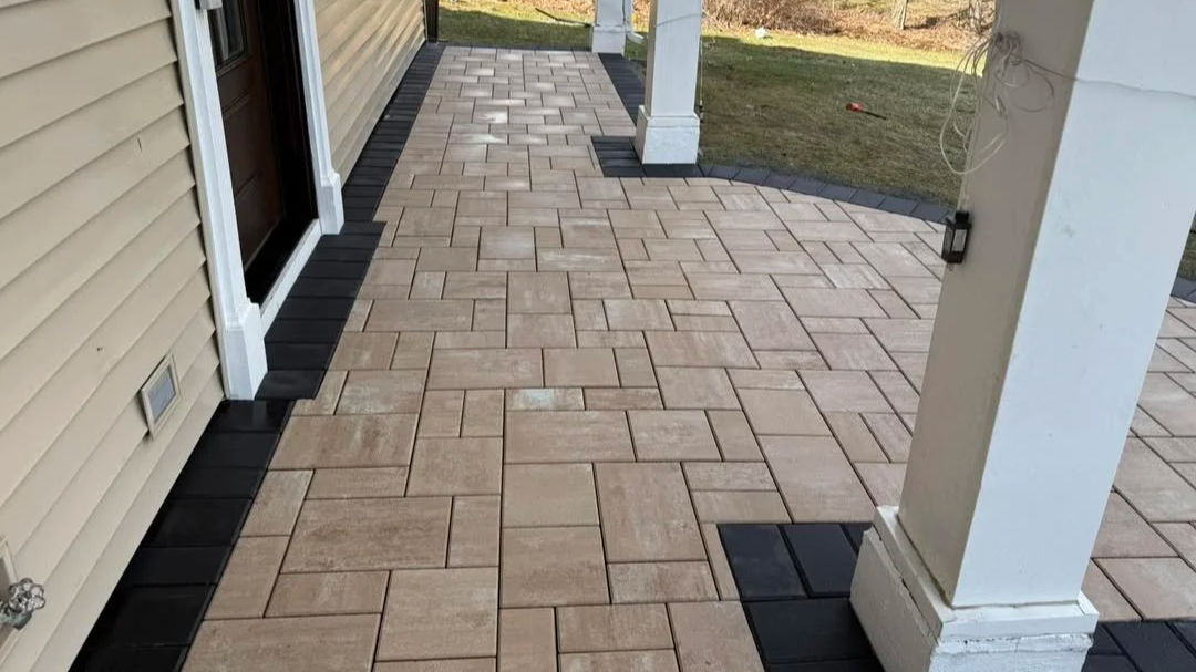 A backyard patio with interlocking rectangular beige paving stones bordered by black tiles, next to a house with beige siding and white trim, with a white column and some exposed wires in the background.