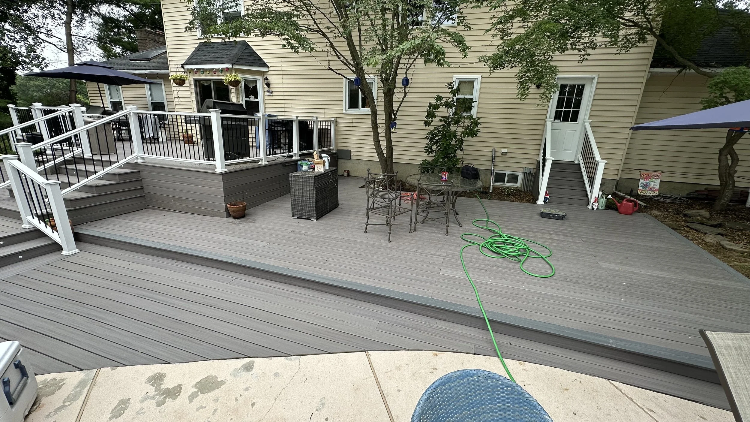A backyard patio with wooden decking, a small table with four chairs, and stairs leading down from a house with beige siding. There are trees and plants around, and various outdoor items are visible.