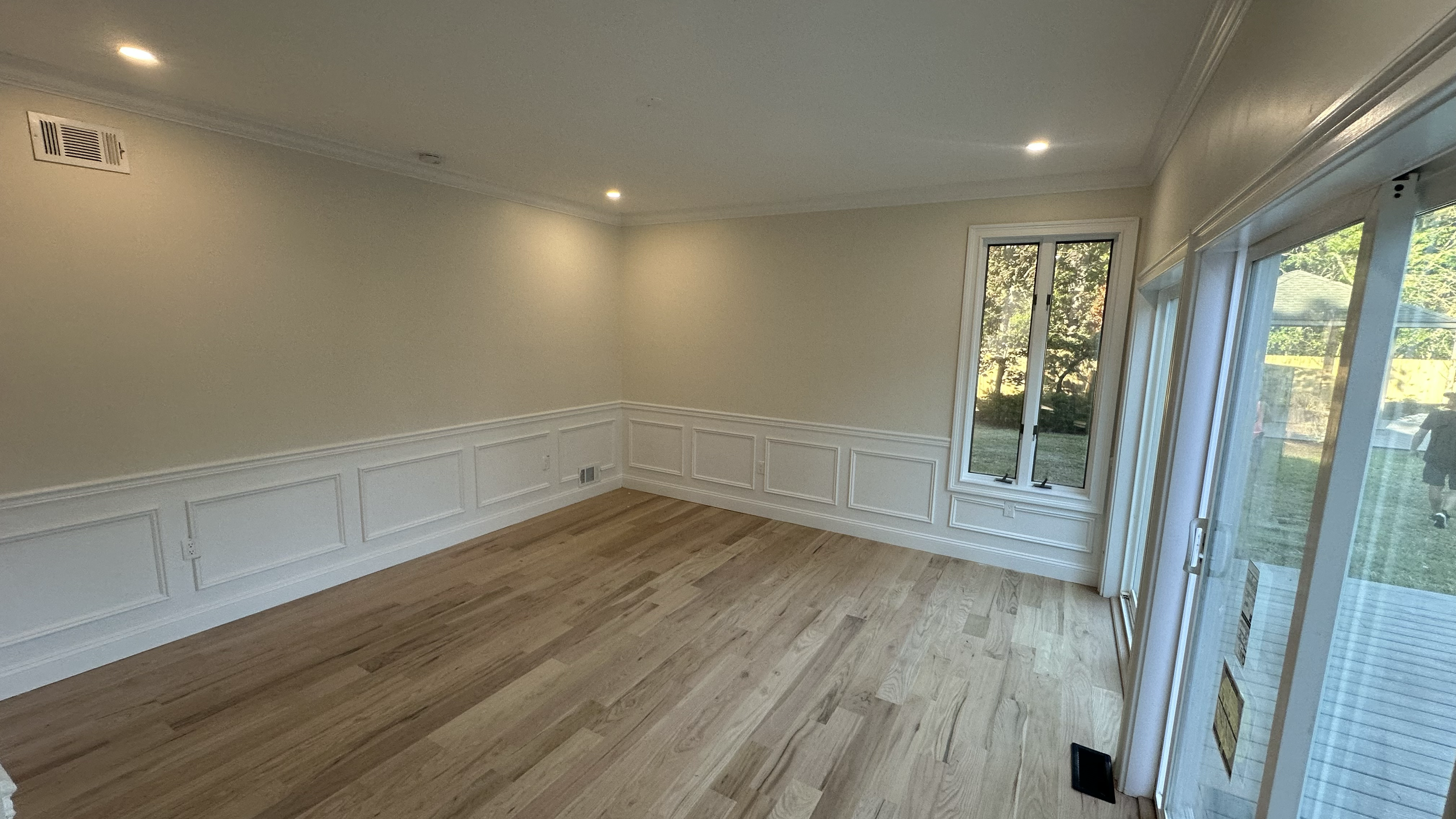 Empty room with hardwood floors, beige walls, white trim, sliding glass door, and windows, with outdoor view of trees and patio.