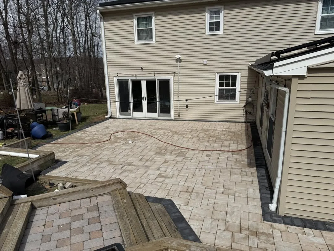 A backyard patio with beige pavers, adjacent to a house with beige siding, and a sliding glass door leading inside. There is a string of lights hanging across the backyard.