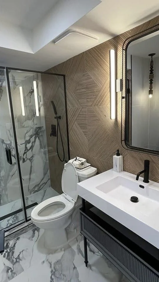 Modern bathroom with a glass-enclosed shower with marble walls, a white toilet, and a white sink with a black faucet. There are stylish vertical light fixtures and a large mirror on a wooden accent wall.
