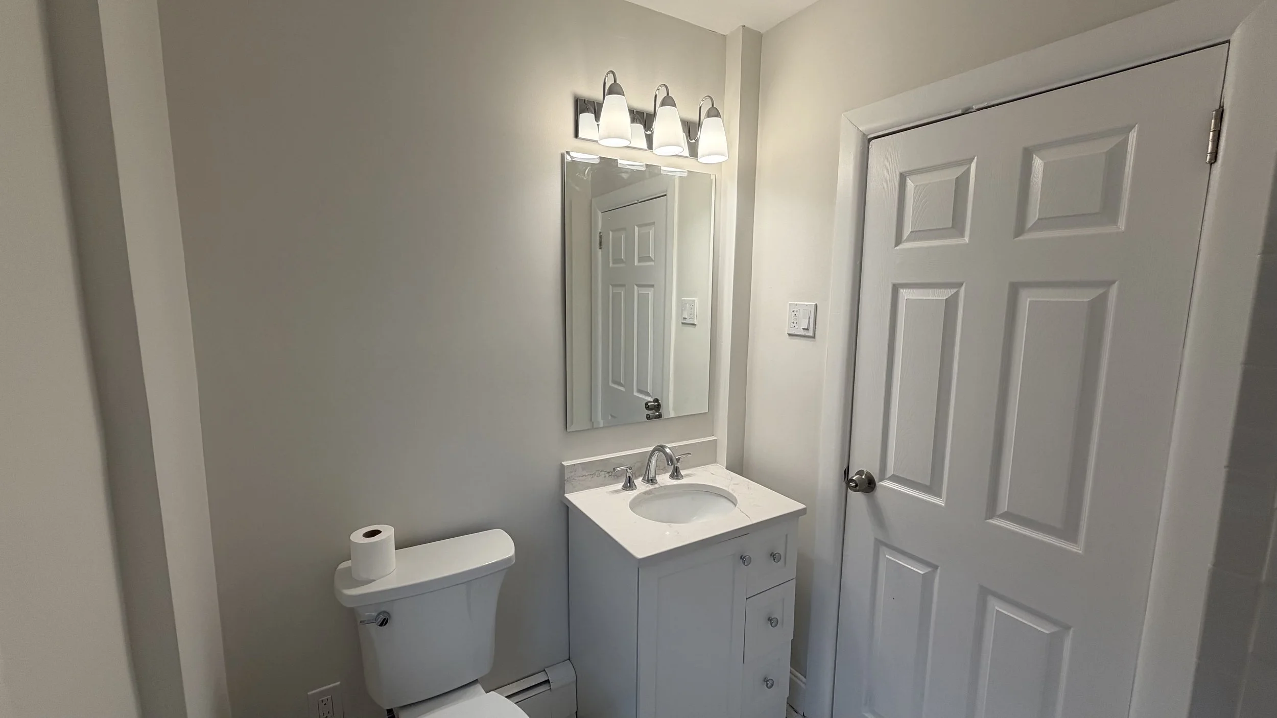 A small bathroom with a white toilet and a white vanity with a sink, mirror, and overhead light fixture, with a closed door on the right side.