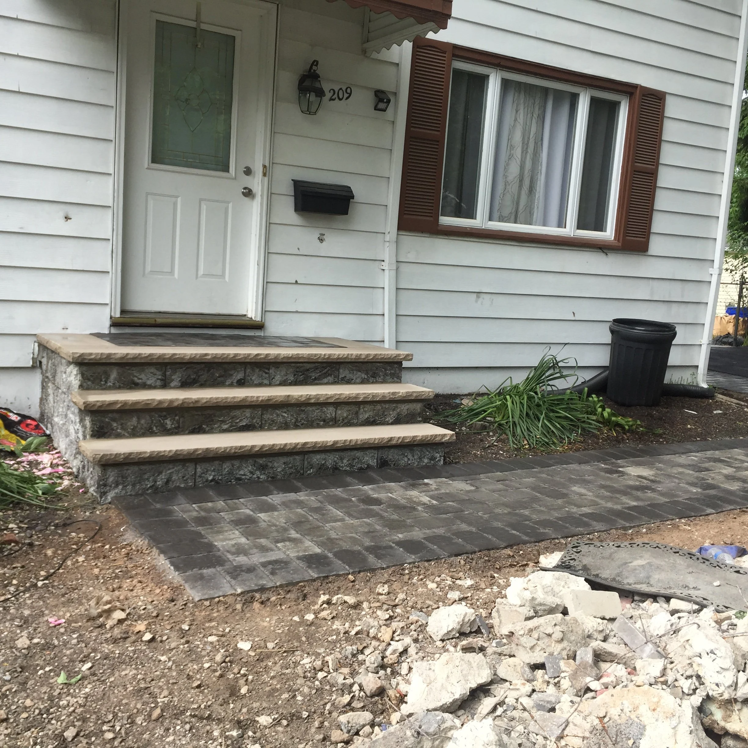 A house with a white front door, a black mailbox, and a window with brown shutters. There are stone steps leading up to the door and a freshly paved brick walkway in front, with some construction debris and dirt nearby.