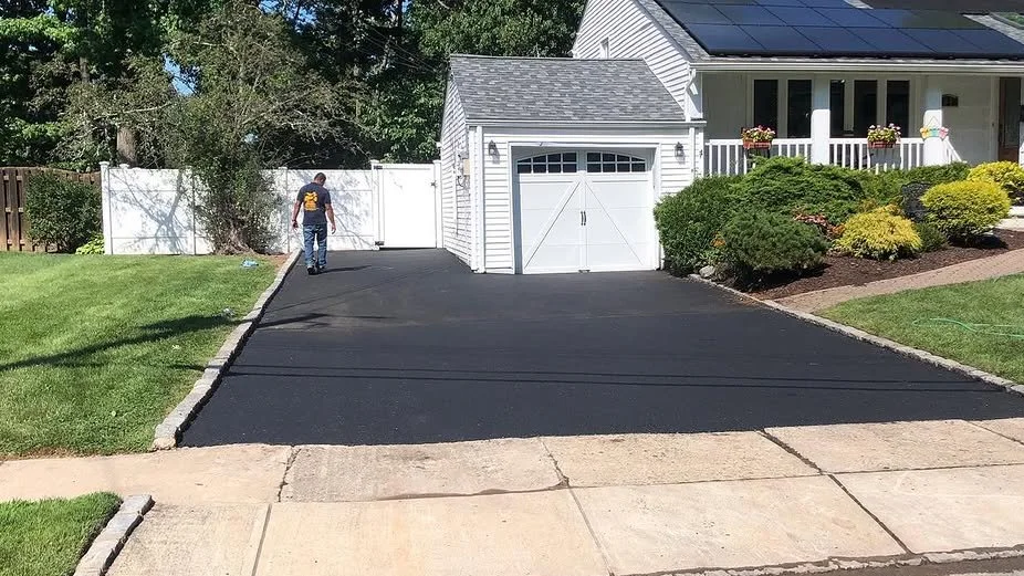 A man is walking on a freshly paved black asphalt driveway in front of a white garage and house, with green lawns and landscaped bushes to the sides.