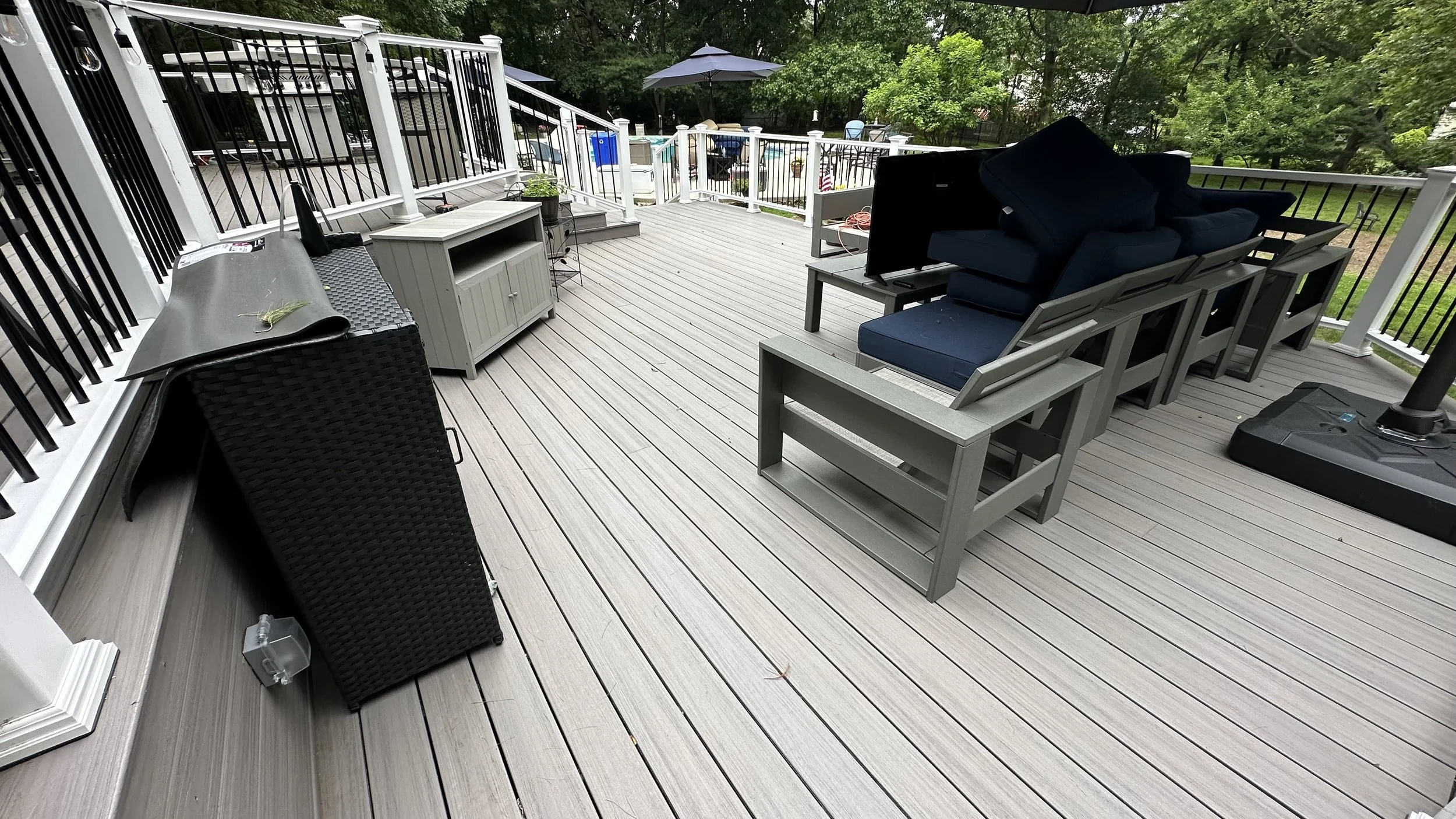 Outdoor deck with furniture including a black wicker storage box, a gray cabinet, dark blue cushioned chairs, a small table, and an umbrella, surrounded by green trees.