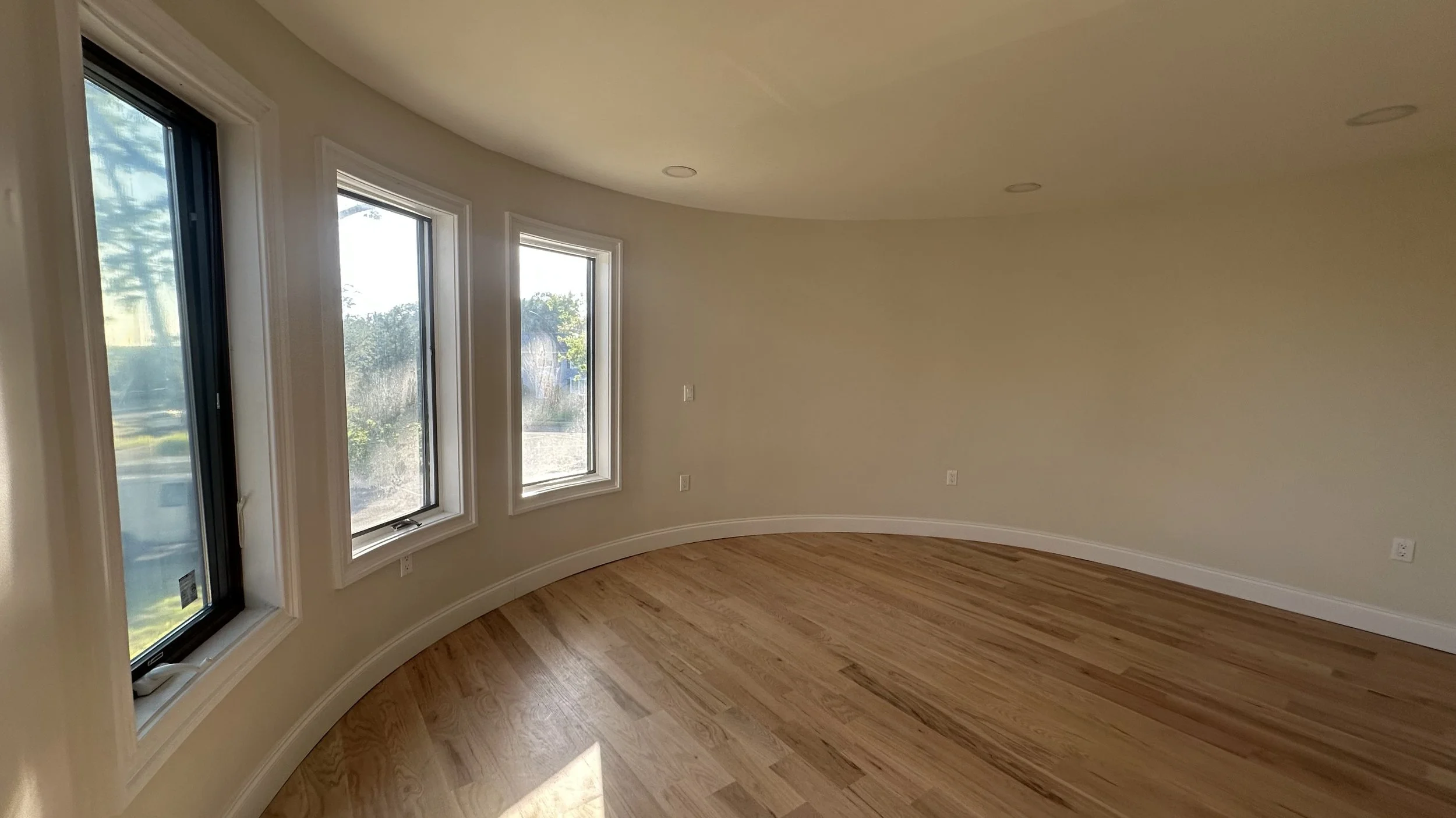 Empty room with three large windows, beige walls, floor made of light wood, and white baseboards.