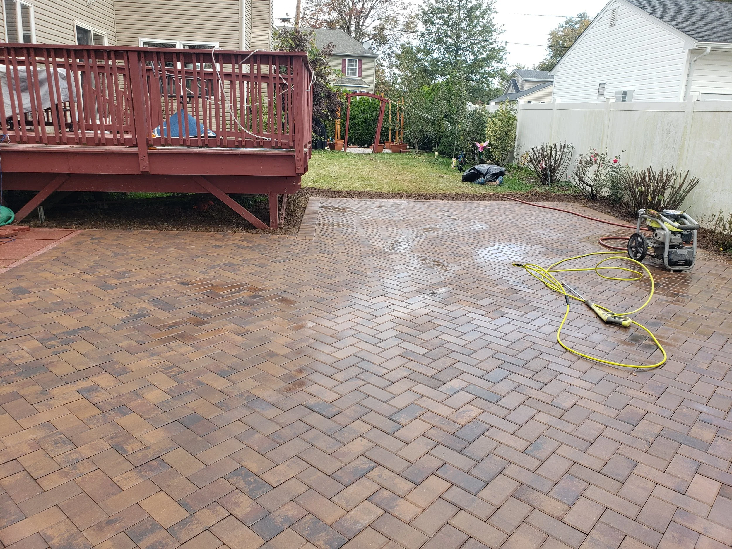 Backyard patio with a newly power-washed brick surface, a red wooden deck, garden beds with bushes and trees, an electric power washer, and garden hoses.