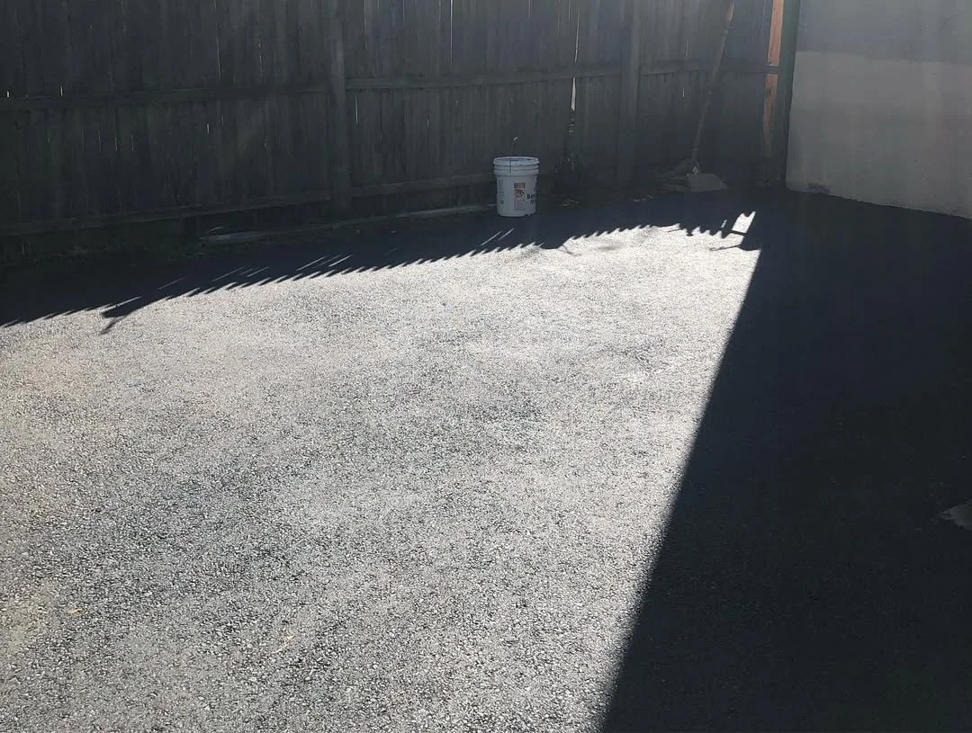 Empty gravel backyard with a wooden fence, a white bucket, and sunlight casting shadows.