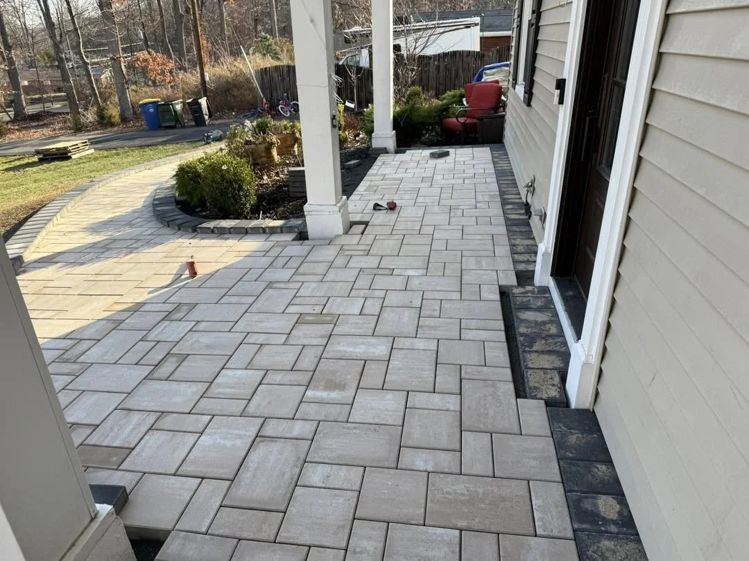 View of a newly paved outdoor patio with beige interlocking bricks, bordered by darker bricks, next to a house with light yellow siding and a door.