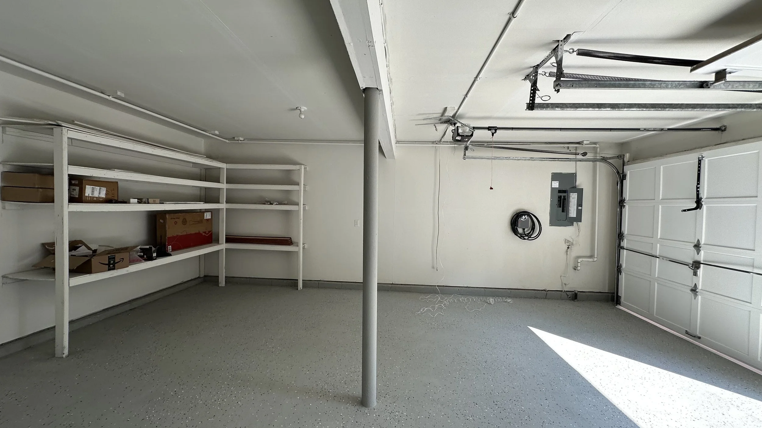 Empty garage with white shelves on the left, a grey garage door on the right, and garage equipment on the wall including an electrical panel and a hose reel.