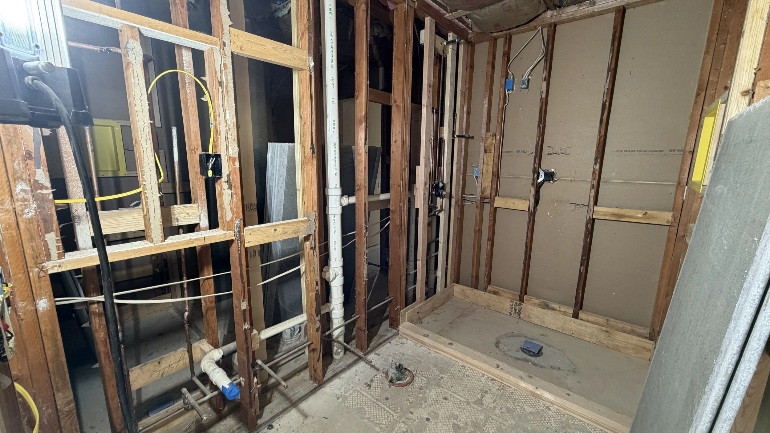 Interior of a bathroom under renovation, showing exposed wooden framing, plumbing pipes, electrical wires, and drywall installed on some walls.