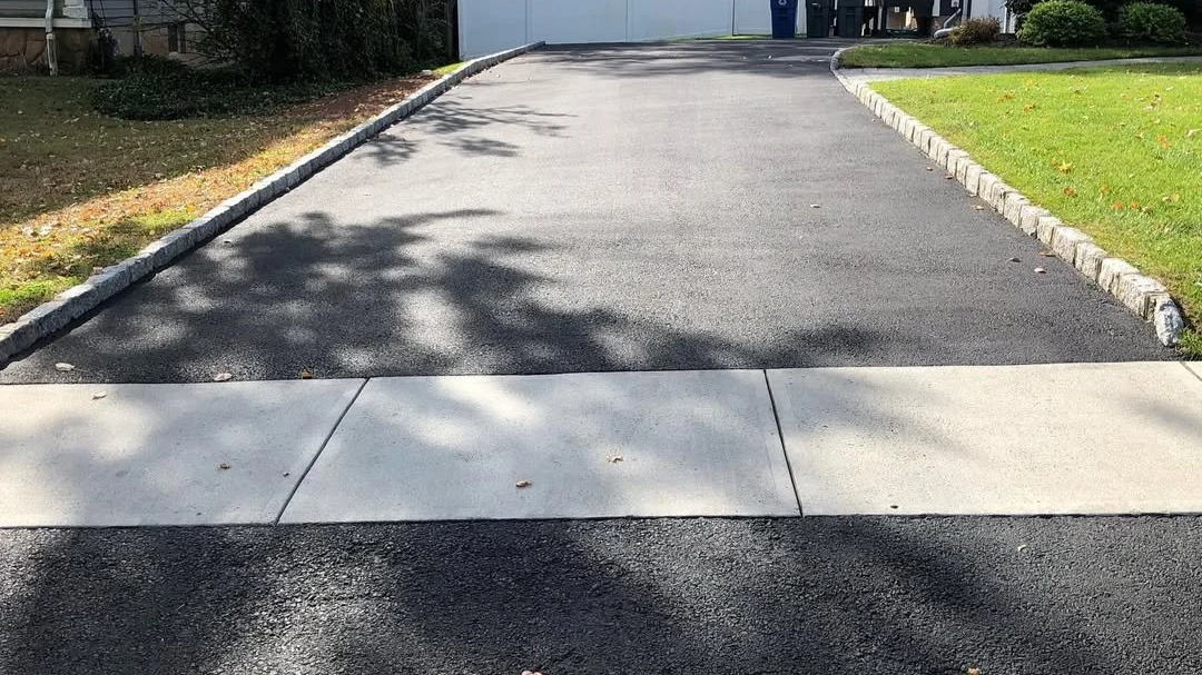 A freshly paved driveway with a concrete sidewalk in the foreground, bordered by lawn on both sides, with shadows of trees cast across the pavement.