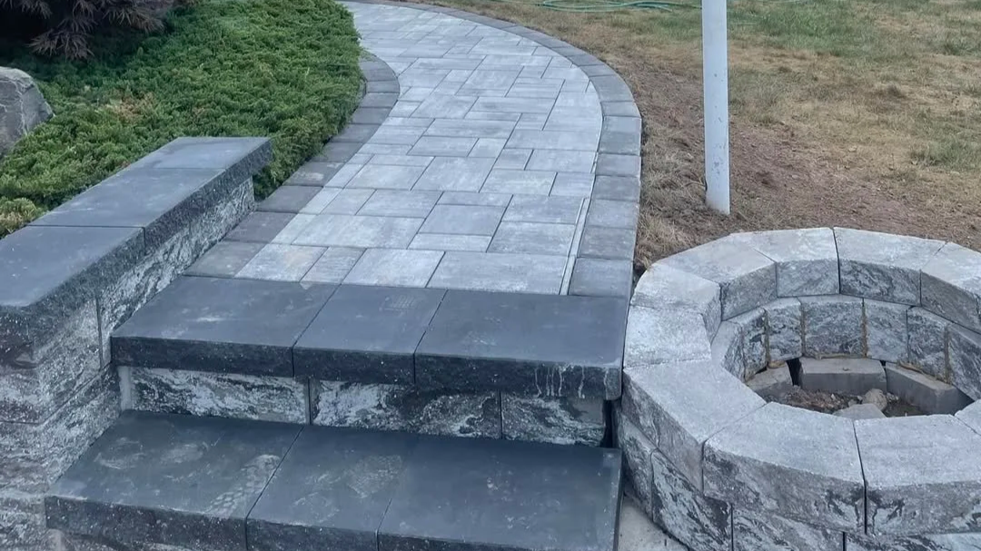 A paved pathway with a path of light-colored rectangular bricks, edged with darker bricks, next to a circular stone fire pit, and surrounded by grassy and landscaped areas.