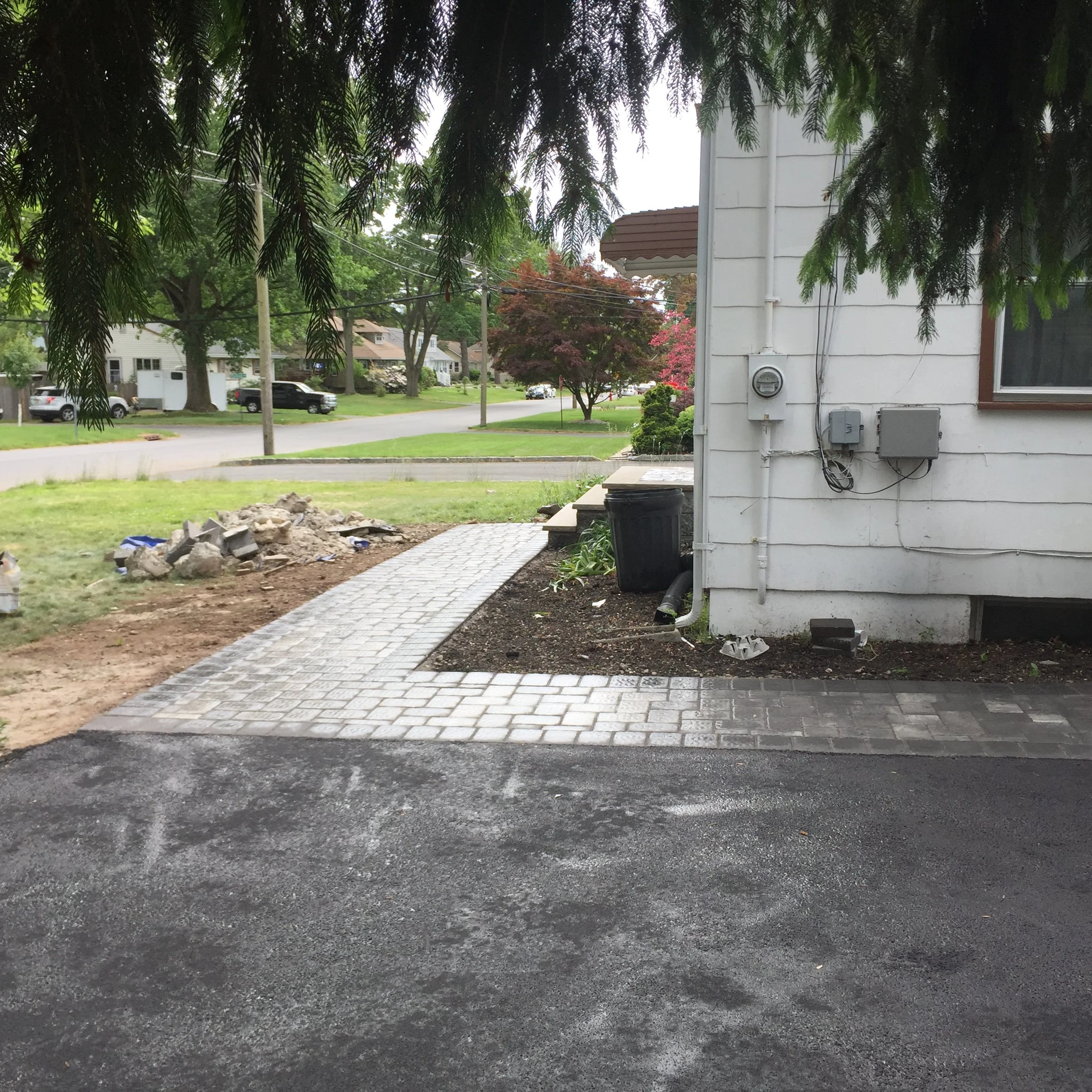A newly paved sidewalk curves around the side of a white house, with a black trash bin and some plants along the house wall. The sidewalk leads to a lawn with a pile of rocks and dirt, under trees with green leaves. In the background, a residential s