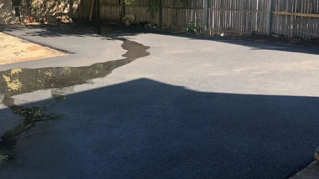 Freshly poured asphalt cooling and drying in a backyard area, bordered by a wooden fence.