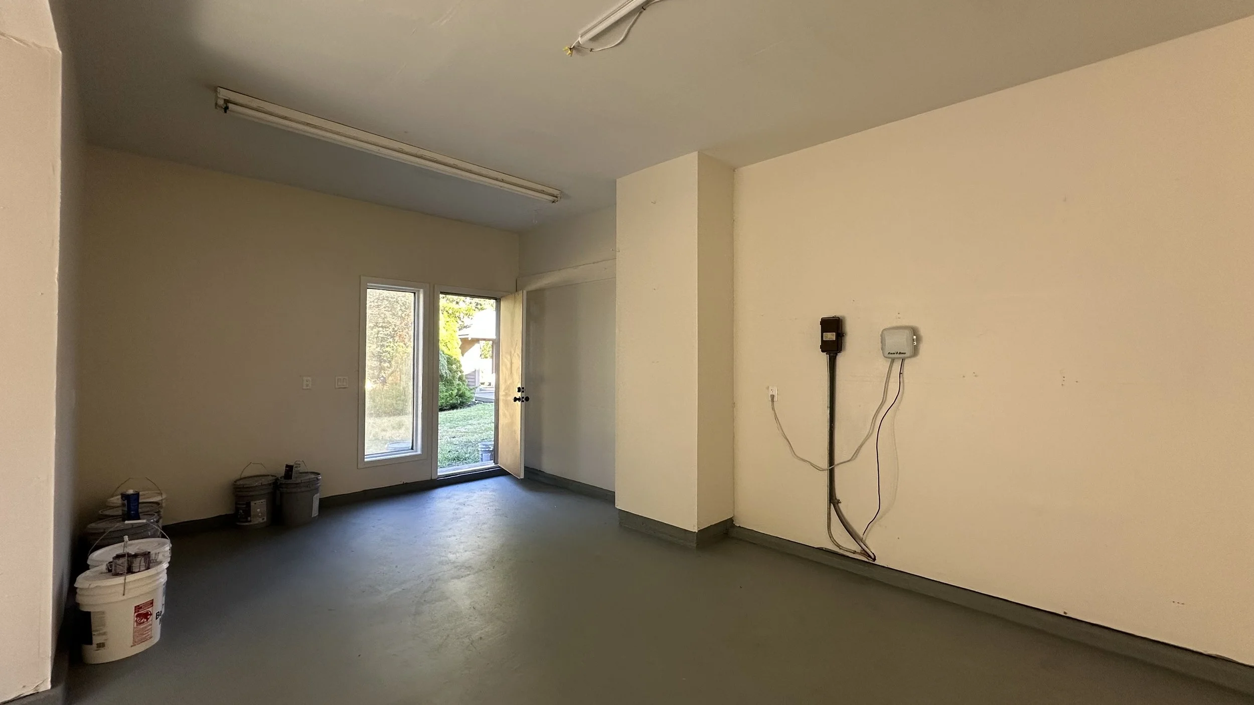 Interior of a room under renovation with beige walls, a gray floor, several buckets of paint in the corner, a window, and a door leading outside. Visible electrical wiring and outlets are on the walls.