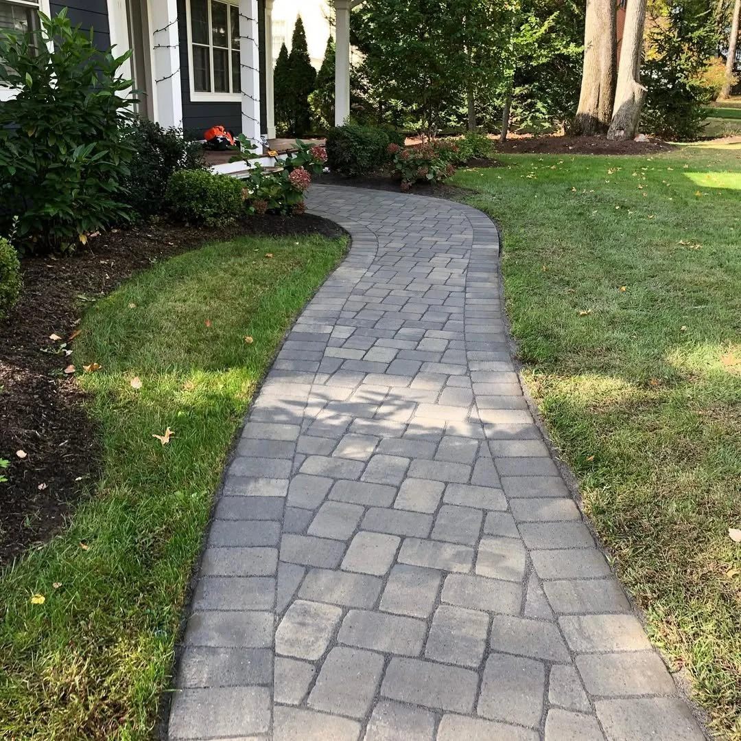 A winding concrete or stone pathway in a garden, leading to a house with a porch, surrounded by green grass, bushes, and trees.