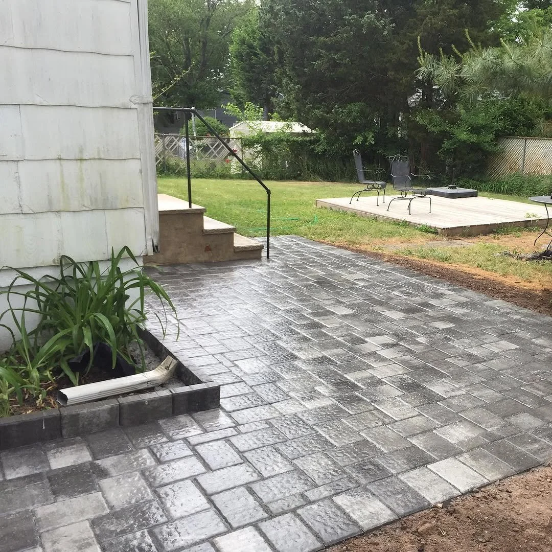 A newly paved gray brick patio extends from a white house with a small staircase and black metal handrail. The patio borders a grassy yard with a wooden deck, outdoor chairs, and a table, surrounded by green trees and bushes.