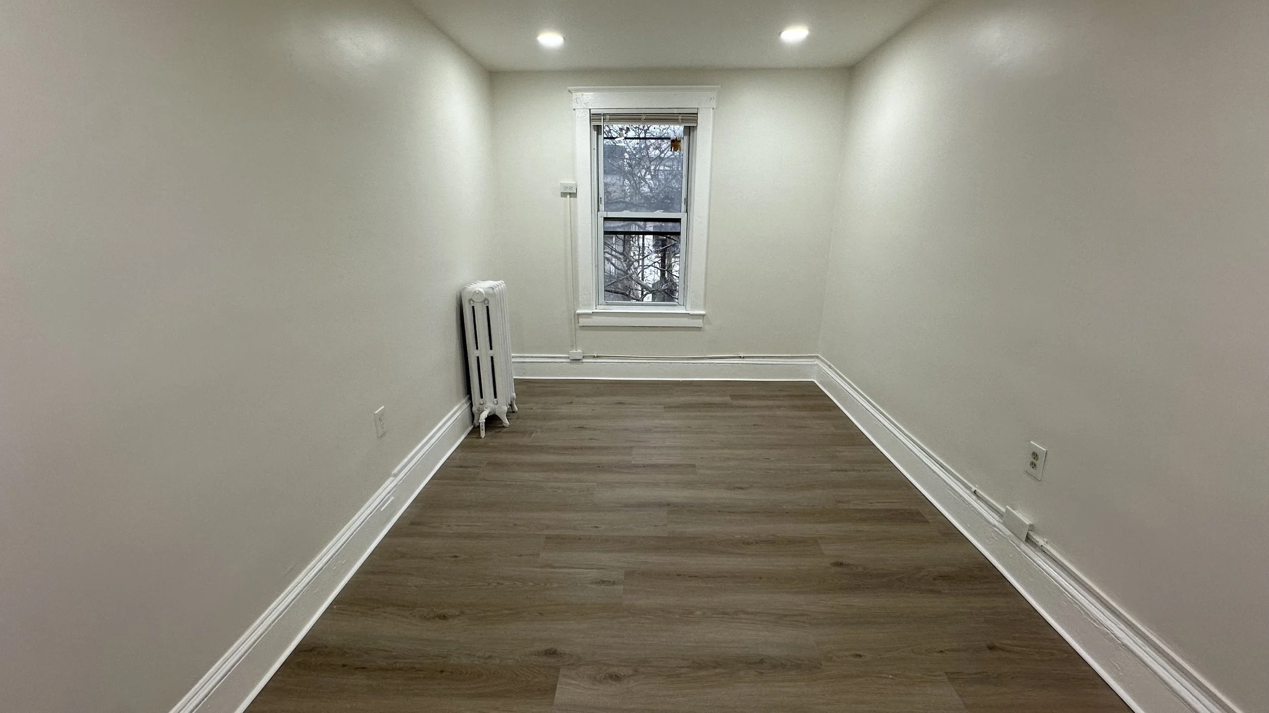Empty room with white walls, wooden floor, a window showing trees outside, white trim, radiator, and electrical outlets.